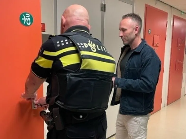Verpleegkundige Daryl staat bij een deur van een politiecel met een politieagent. 