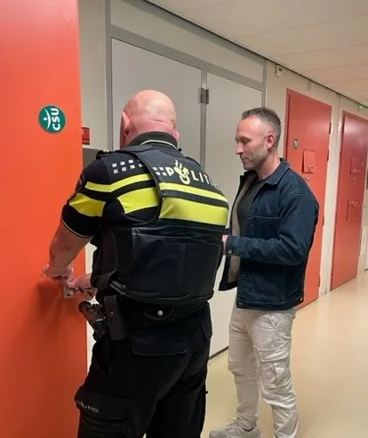 Verpleegkundige Daryl staat bij een deur van een politiecel met een politieagent. 