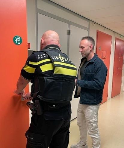 Verpleegkundige Daryl staat bij een deur van een politiecel met een politieagent. 